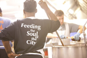 A volunteer leans on a ladle in a soup bowl, facing away from the camera. On the back of their sweatshirt is written "Feeding the Soul of the City"