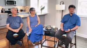 Ilan Rosenblit sits at left, his wife Merav sits in the middle, and on the right sits Cebastian Hilton. All are wearing blue and sitting in a living room of a house with the kitchen visible behind them.