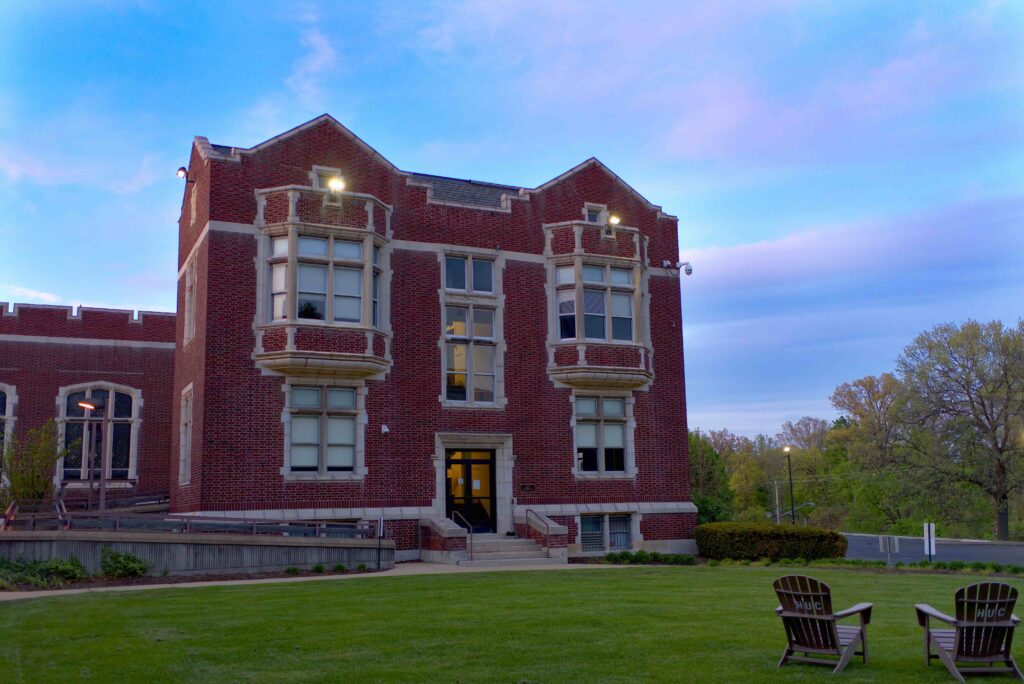 A brick building covered in windows with a lawn in front and a sky above. The sky is just barely starting to turn red as the sun sets to the left of the building.