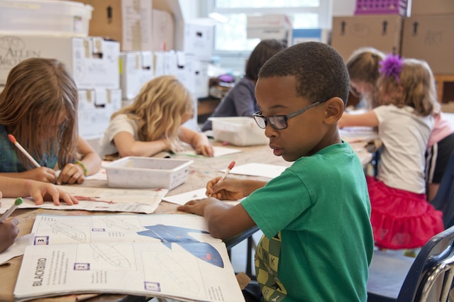 A little boy in glasses and a green shirt holds a pencil while drawing and referencing a book with schematics of planes.