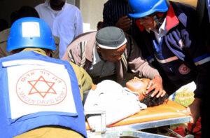 An Israeli woman lies on a stretcher after being injured from a rocket attack from Gaza while medics from Magen David Adom surround her. A man leans over her with his hand on her head.