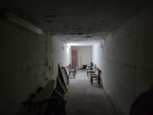 A photo of an Israeli bomb shelter that looks like a wide, white concrete hallway with a light at the far end and chairs lining the sides.