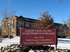 A red sign with white text with a building and trees behind the sign. The sign reads "Hebrew Union College-Jewish Institute of Religion. Klau Library. Skirball Museum Cincinnati Branch. Marcus Center - American Jewish Archives"