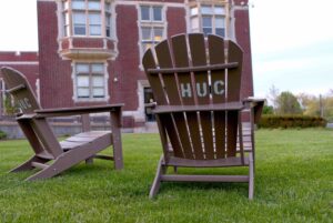 Two adirondack lawn chairs on a green lawn, with a building in the background with red brick. One of the chairs has its back facing the camera, on which there are painted letters that say "HUC" in reference to the Hebrew Union College-Jewish Institute of Religion.