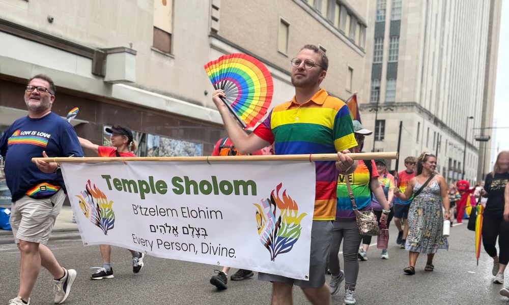 two men hold a banner that reads "Temple Sholom, Btzelm Eloheim, in English Transliteration, and Every Person is Divine in English" One of the two man fans himself with a rainbow fan