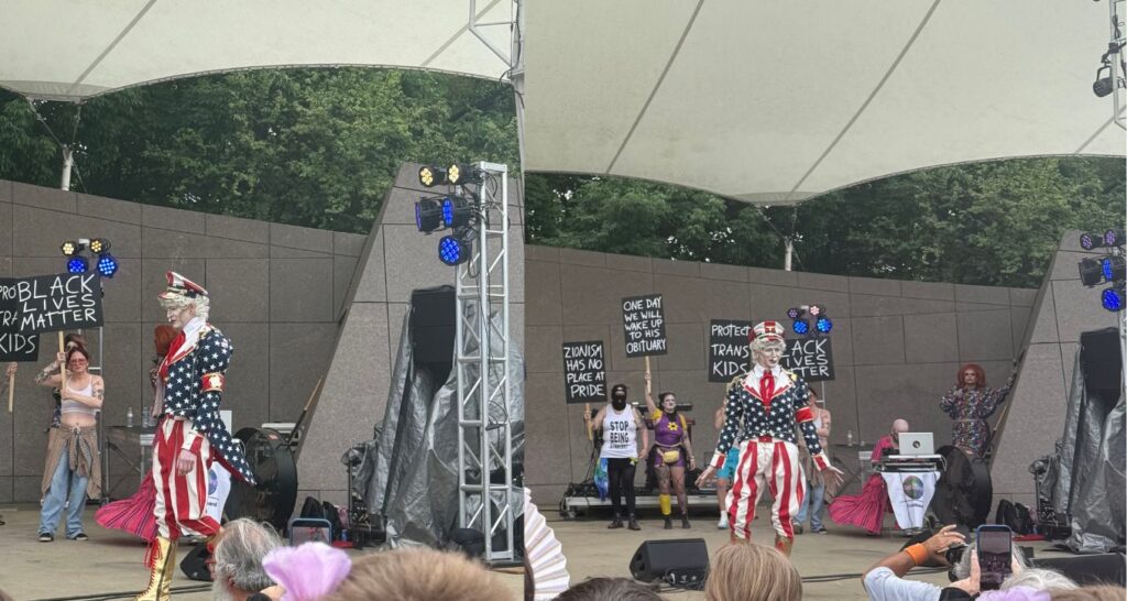 A drag performer on stage dressed in a costume that blends Hitler with Uncle Sam. Surrounded by people holding signs that read progressive political slogans, and one antizionist sign